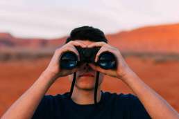Man facing camera looking through binoculars with desert in background behind him