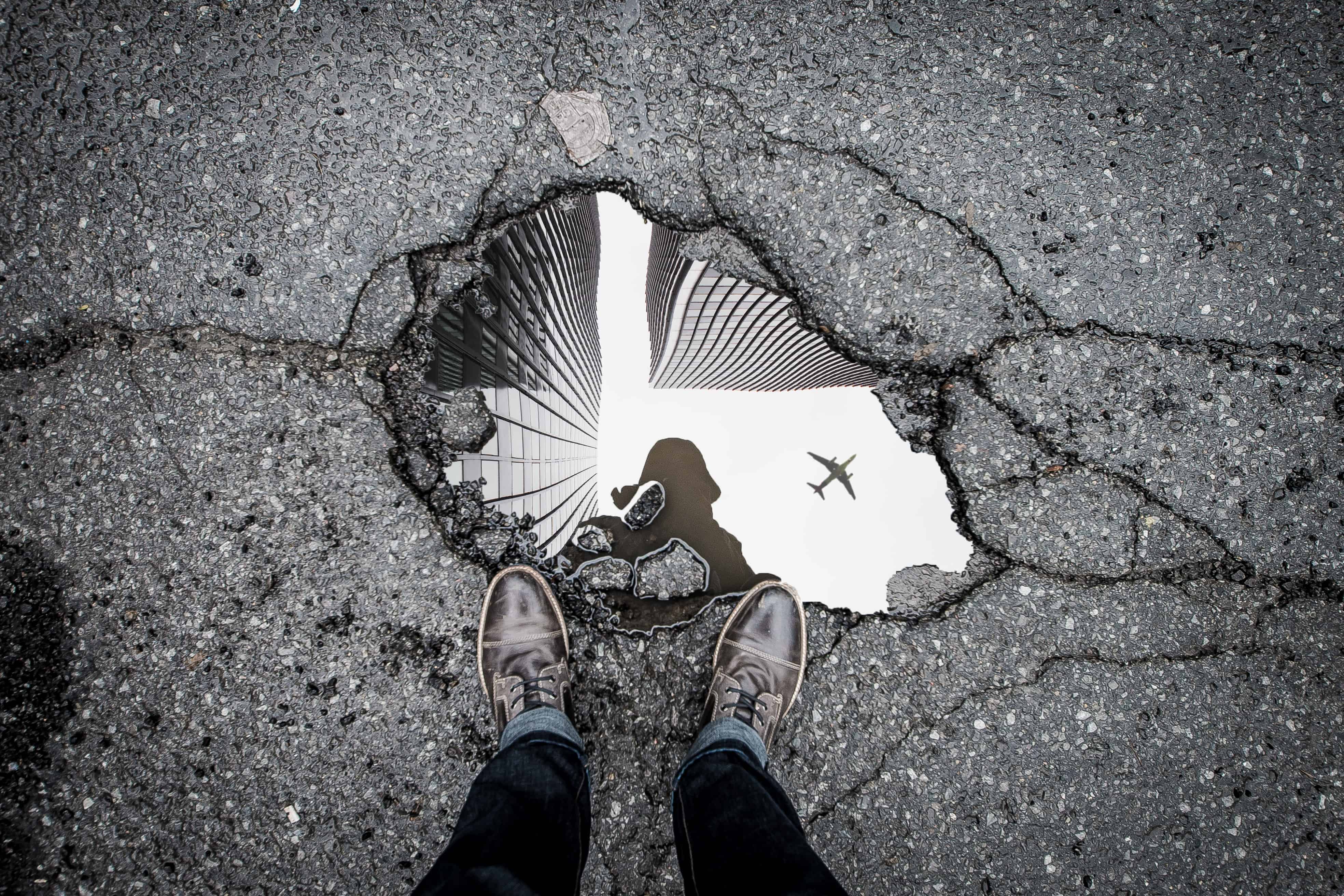 Feet and puddle with puddle reflecting a person, buildings, and a plane flying overhead