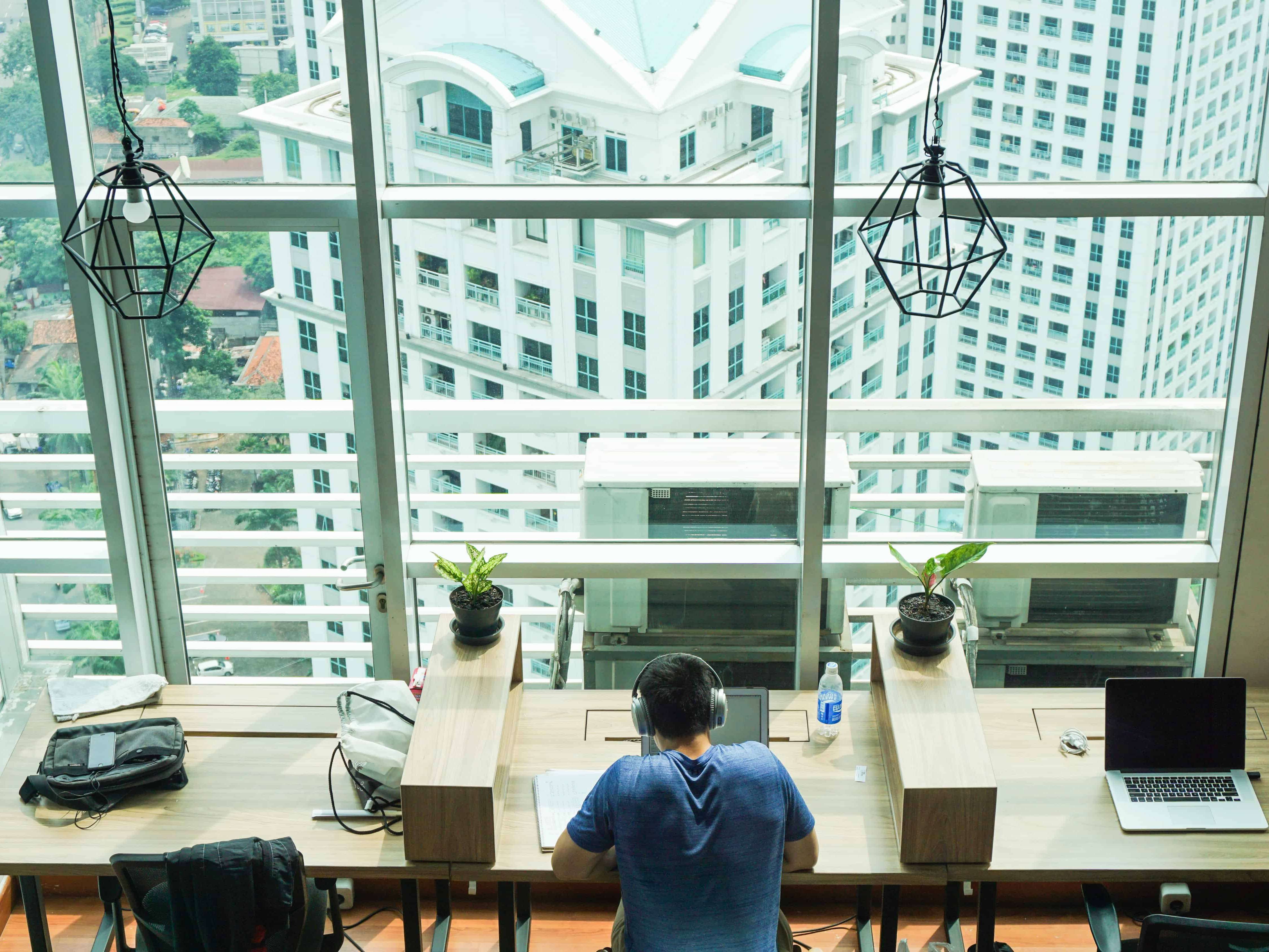 Man doing work on computer at wooden desk in front of glass windows overlooking cityscape