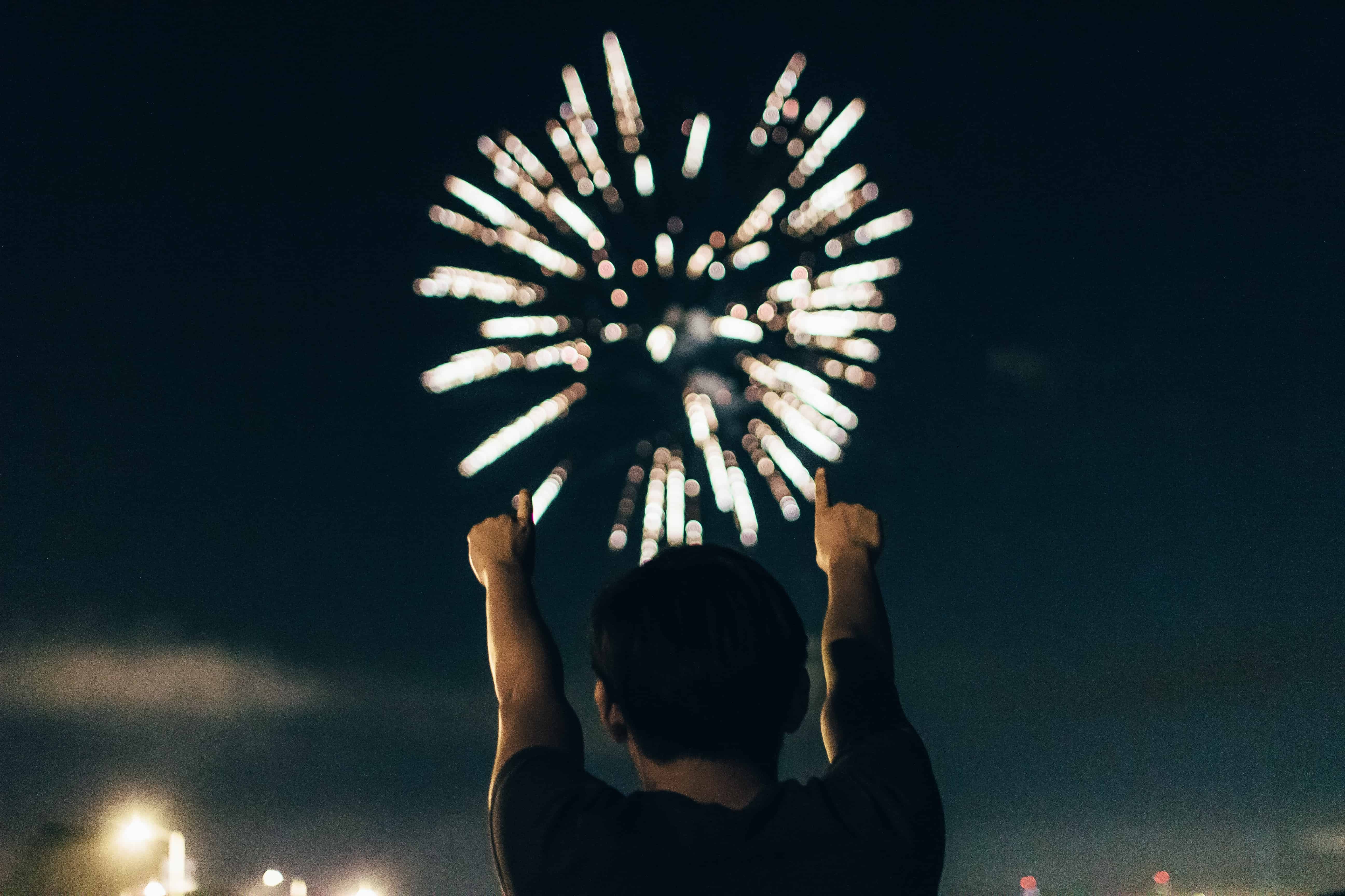 Photo of man facing away from camera with arms up pointing at firework exploding in night sky