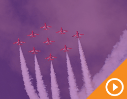Nine planes flying in formation across the sky behind a purple transparency with a white play button on an orange triangle in the bottom right corner