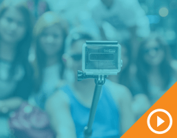 GoPro on selfie stick with blurry crowd in background behind blue transparency with white play button on an orange triangle in the bottom right corner