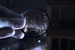 Person holding lens against night city where city in lens is in focus, and surrounding background is out of focus