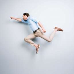 Happy, attractive, handsome, young, cool, joyful man with bristle jumping in air showing superman pose over grey background
