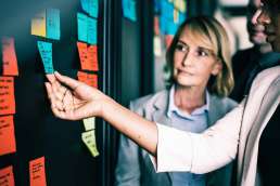 Woman grasping sticky note on wall of with other sticky notes while co-workers look on
