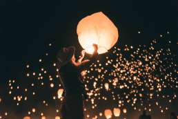 Woman releasing a paper lantern with many other paper lanterns being released into the night sky behind her