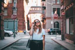 Woman walking down street adjusting sunglasses with brick buildings behind her
